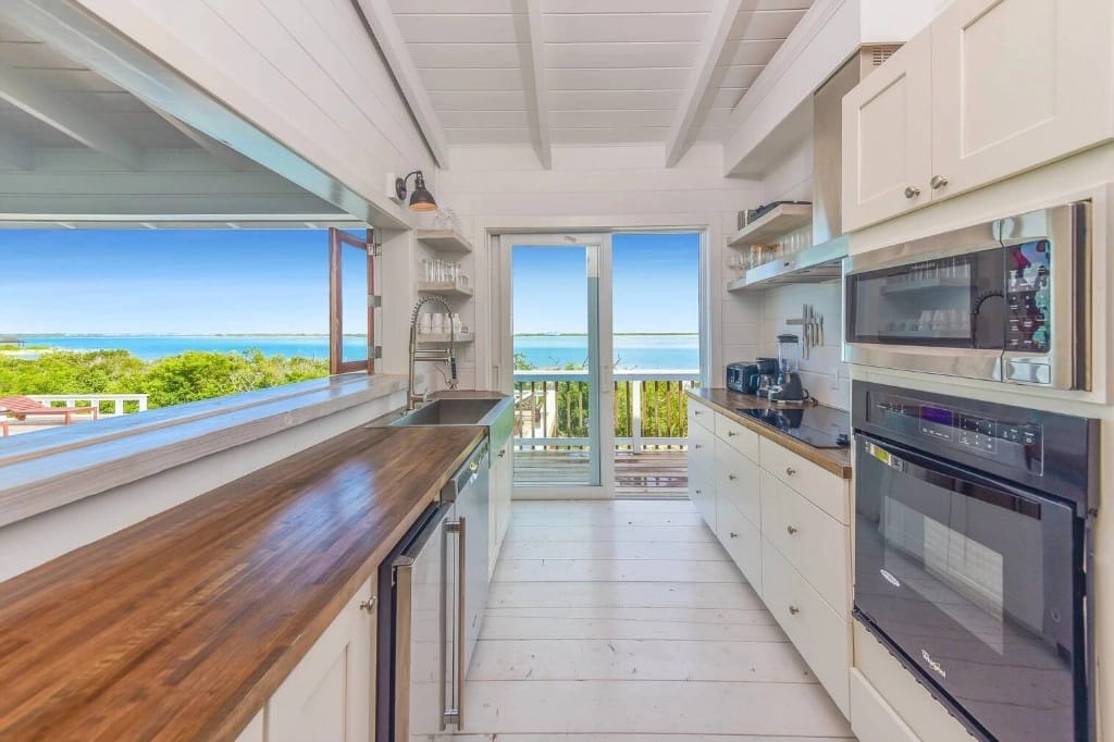 A bright coastal kitchen with white cabinetry, wood countertops, and modern appliances opens to a balcony with sweeping views of turquoise waters and lush greenery.