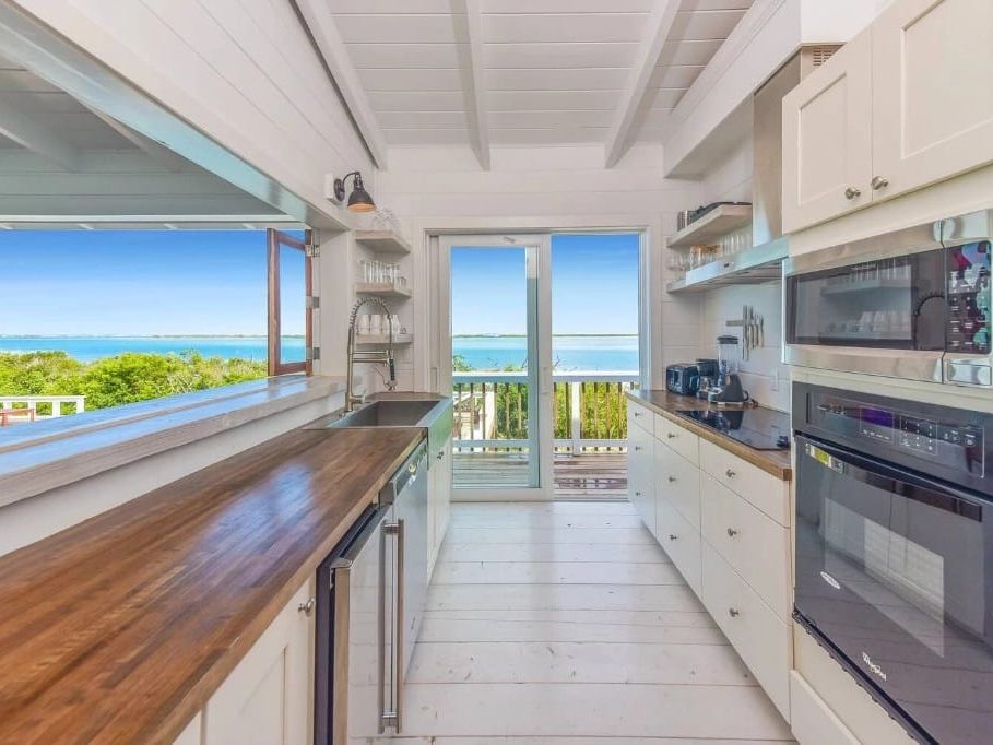 A bright coastal kitchen with white cabinetry, wood countertops, and modern appliances opens to a balcony with sweeping views of turquoise waters and lush greenery.