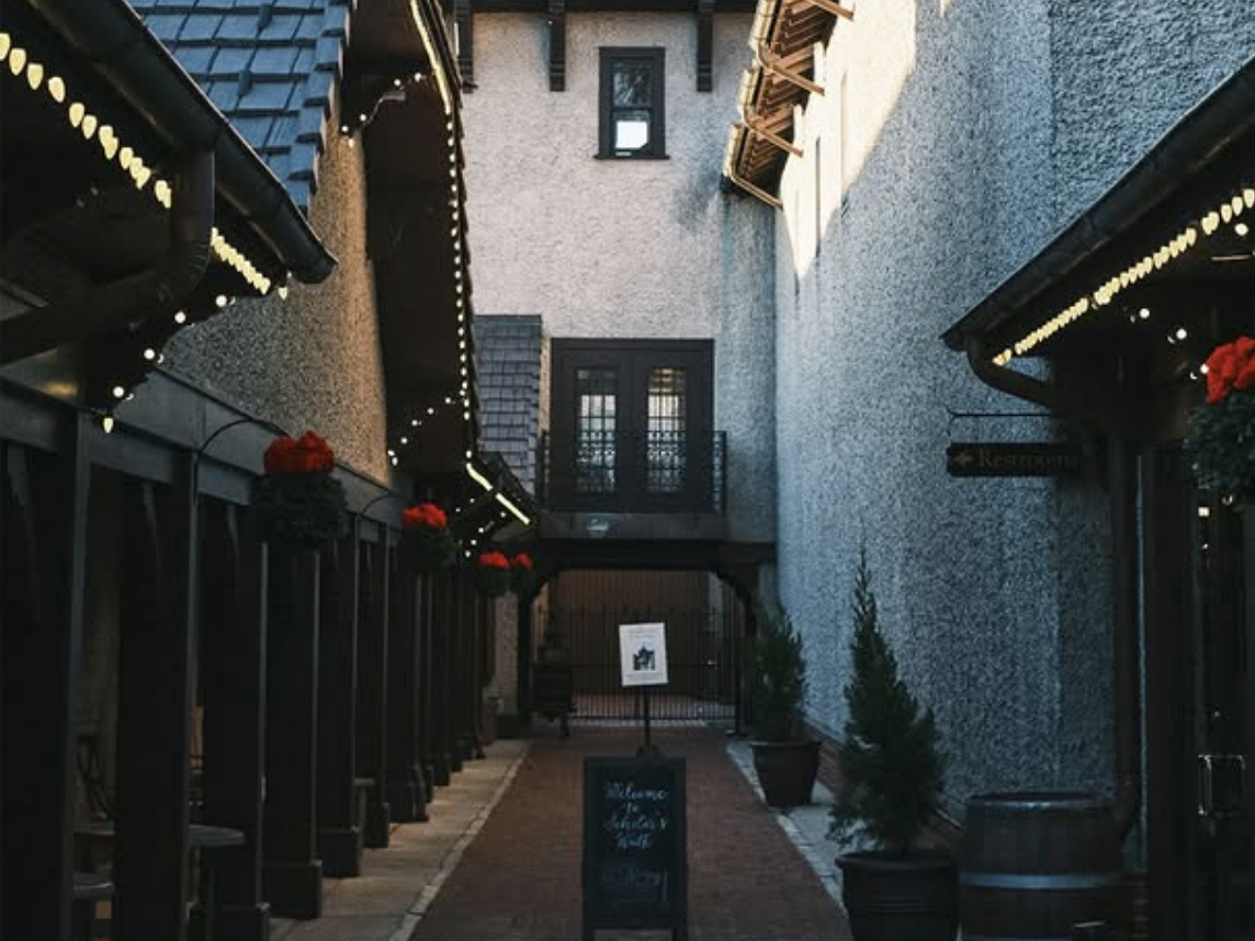 Charming narrow alleyway with stone walls, string lights, potted plants, and a rustic sign leading to a courtyard entrance