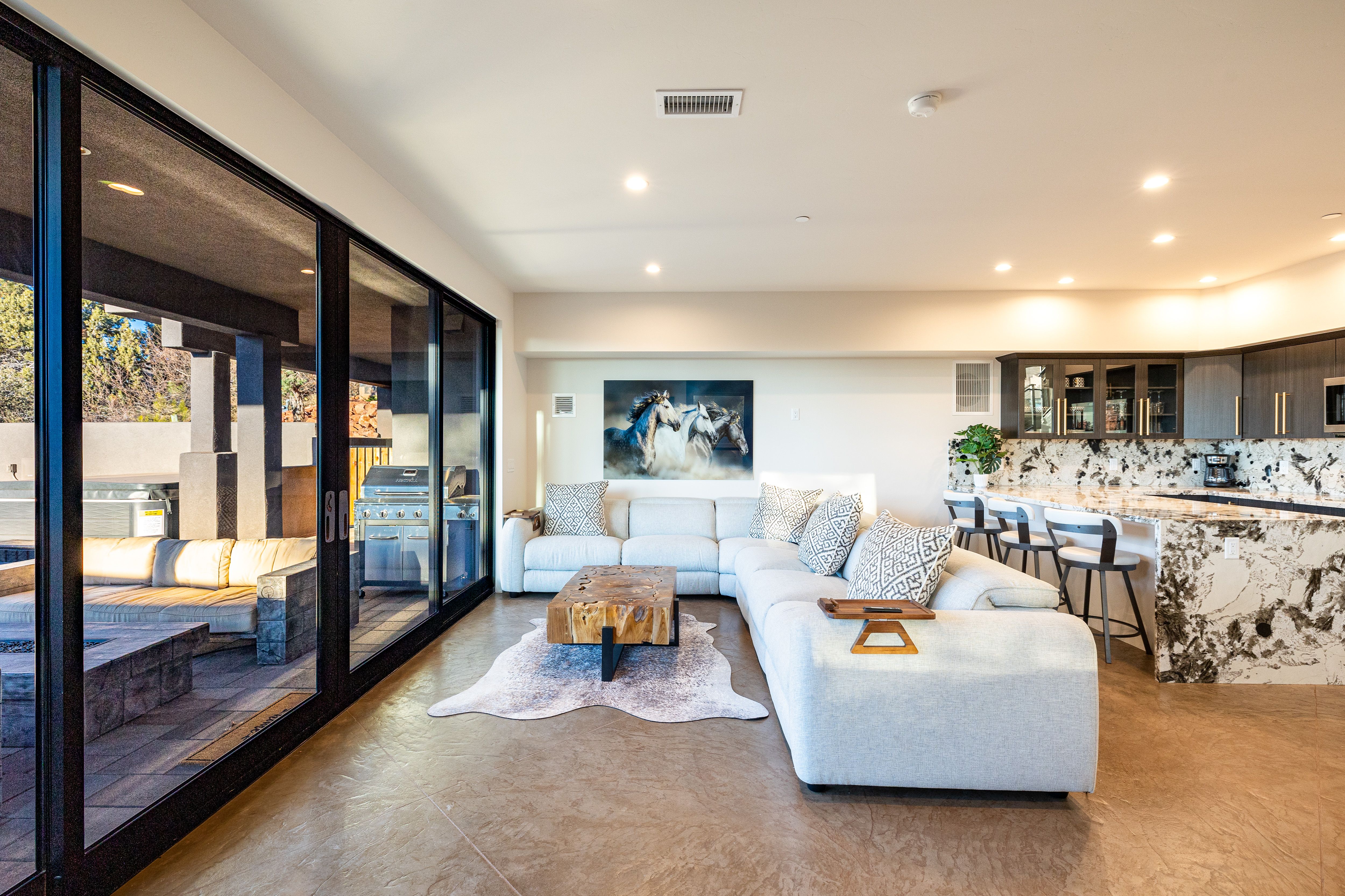 A modern open-concept living room features a light gray sectional sofa, rustic wood coffee table, and large sliding glass doors that open to a covered outdoor lounge with a grill.
