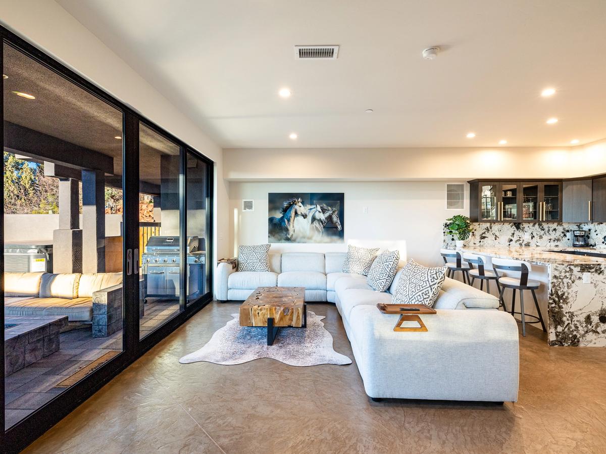A modern open-concept living room features a light gray sectional sofa, rustic wood coffee table, and large sliding glass doors that open to a covered outdoor lounge with a grill.