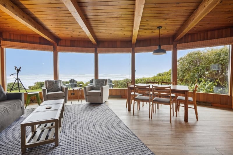 A warm, wood-paneled room with large windows showing a wide view of the ocean. The cozy chairs and simple dining setup make it feel like an easy place to unwind. The sunlight brings out the natural colors of the space.
