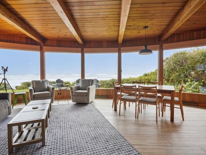 A warm, wood-paneled room with large windows showing a wide view of the ocean. The cozy chairs and simple dining setup make it feel like an easy place to unwind. The sunlight brings out the natural colors of the space.