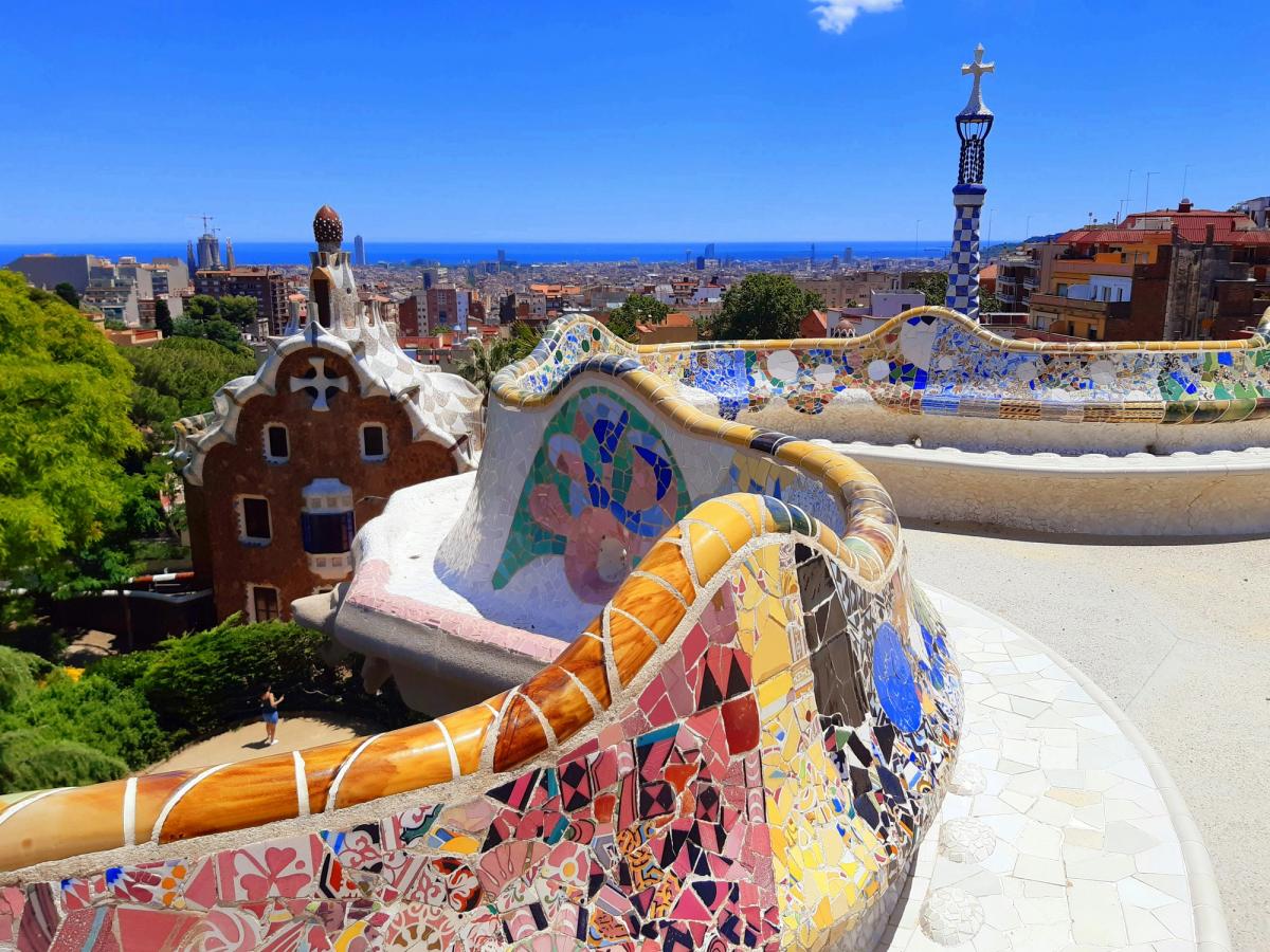 A vibrant, mosaic-tiled serpentine bench in Park Güell overlooks the cityscape of Barcelona, Spain, with whimsical Gaudí architecture and the Mediterranean Sea in the background.