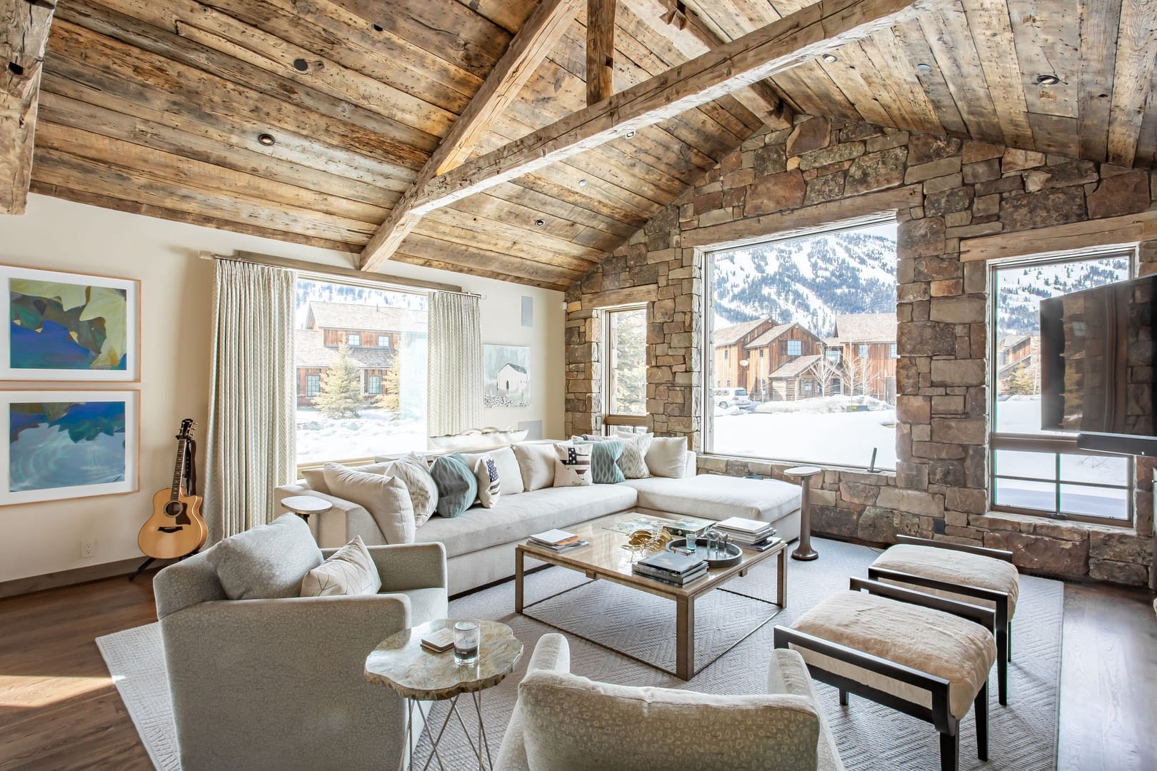 A cozy mountain living room with reclaimed wood beam ceilings, a stone accent wall, plush neutral seating, and large windows framing snowy alpine views.