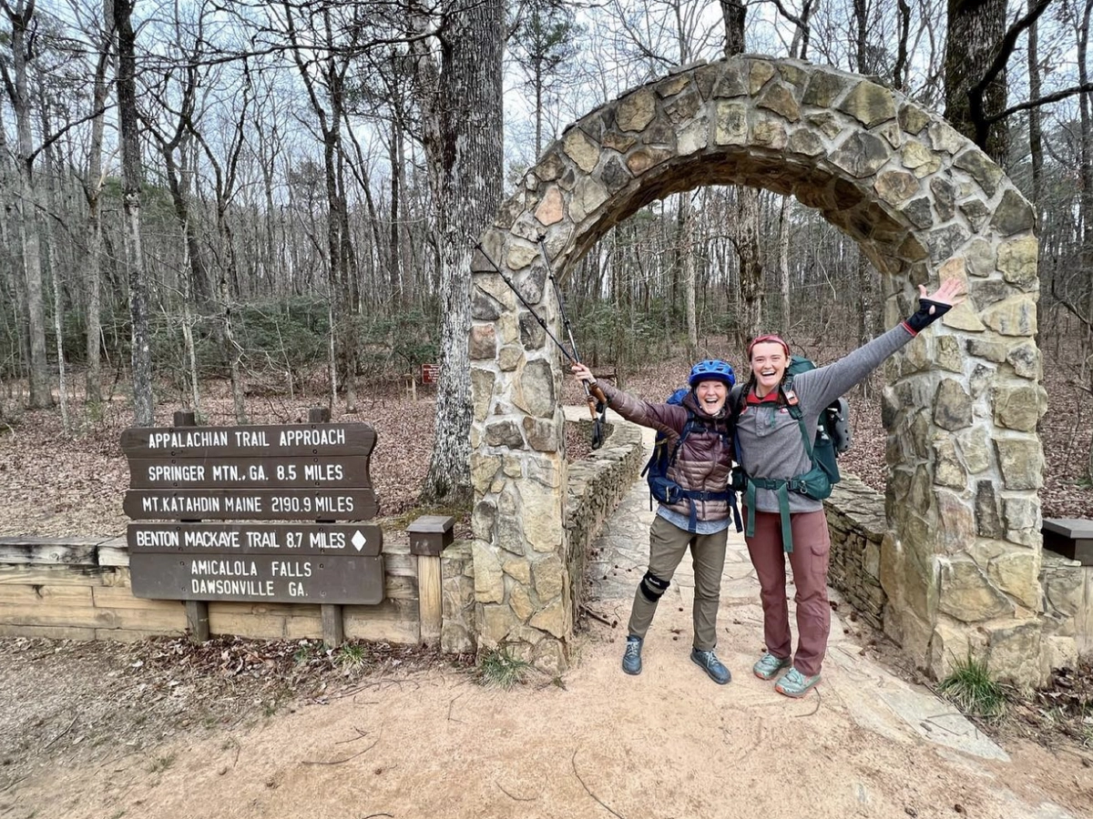 Two joyful hikers pose under the stone arch at the Appalachian Trail Approach in Georgia, beside a sign showing distances to Springer Mountain, Mount Katahdin, and Amicalola Falls.