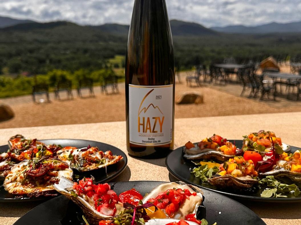 A bottle of Hazy Mountain wine is paired with colorful plates of oysters and fresh toppings overlooking the Blue Ridge Mountains. The stunning view and gourmet pairing showcase Virginia’s elevated mountain wine experience.