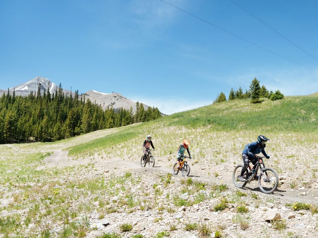 Three mountain bikers ride along a trail through grassy slopes and forested hills in Big Sky. With helmets and full gear, they’re set for a fun ride under the summer sun and scenic mountain views.