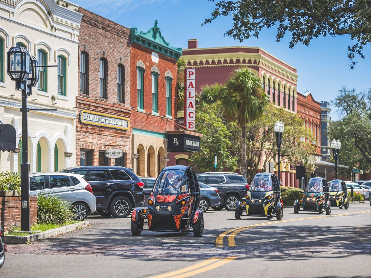 A row of futuristic, three-wheeled Arcimoto electric vehicles cruising through a charming downtown lined with historic brick buildings. The scene is vibrant with palm trees, classic street lamps, and pedestrians enjoying the sunny day