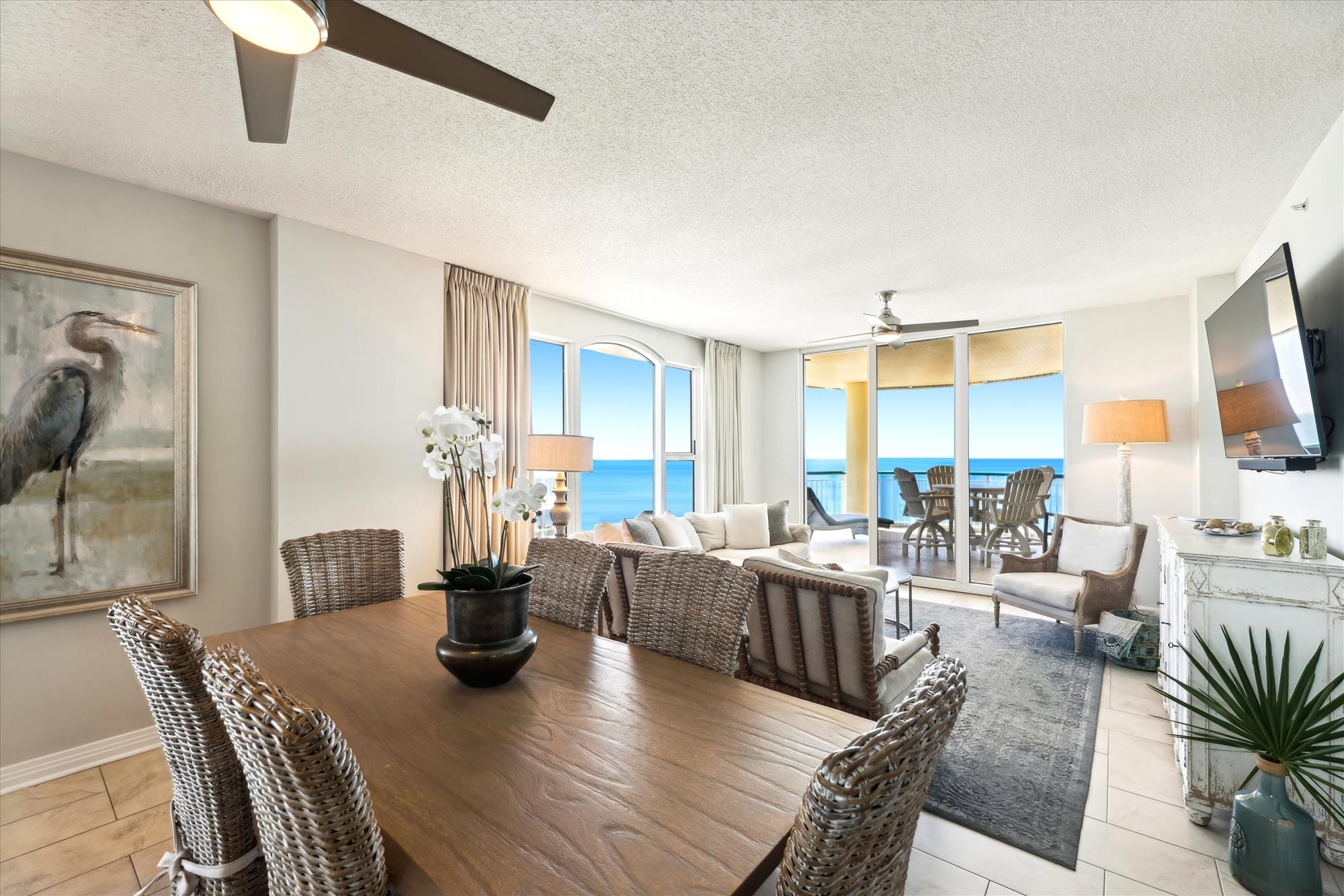 A bright, coastal living room with wicker chairs and soft neutral tones opens to a wide balcony overlooking the Gulf of Mexico. Natural light fills the space, creating a calm beach-house vibe perfect for family gatherings.