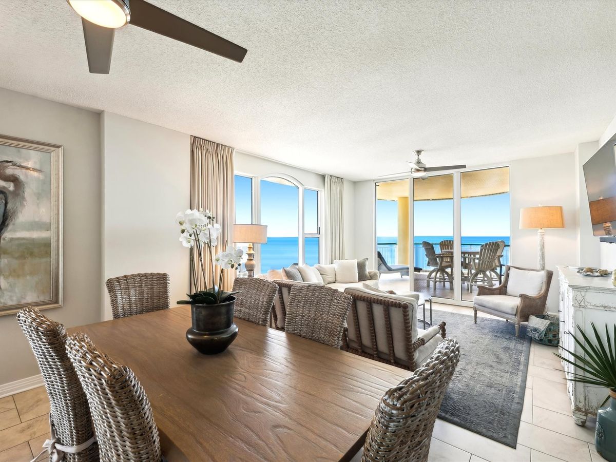 A bright, coastal living room with wicker chairs and soft neutral tones opens to a wide balcony overlooking the Gulf of Mexico. Natural light fills the space, creating a calm beach-house vibe perfect for family gatherings.