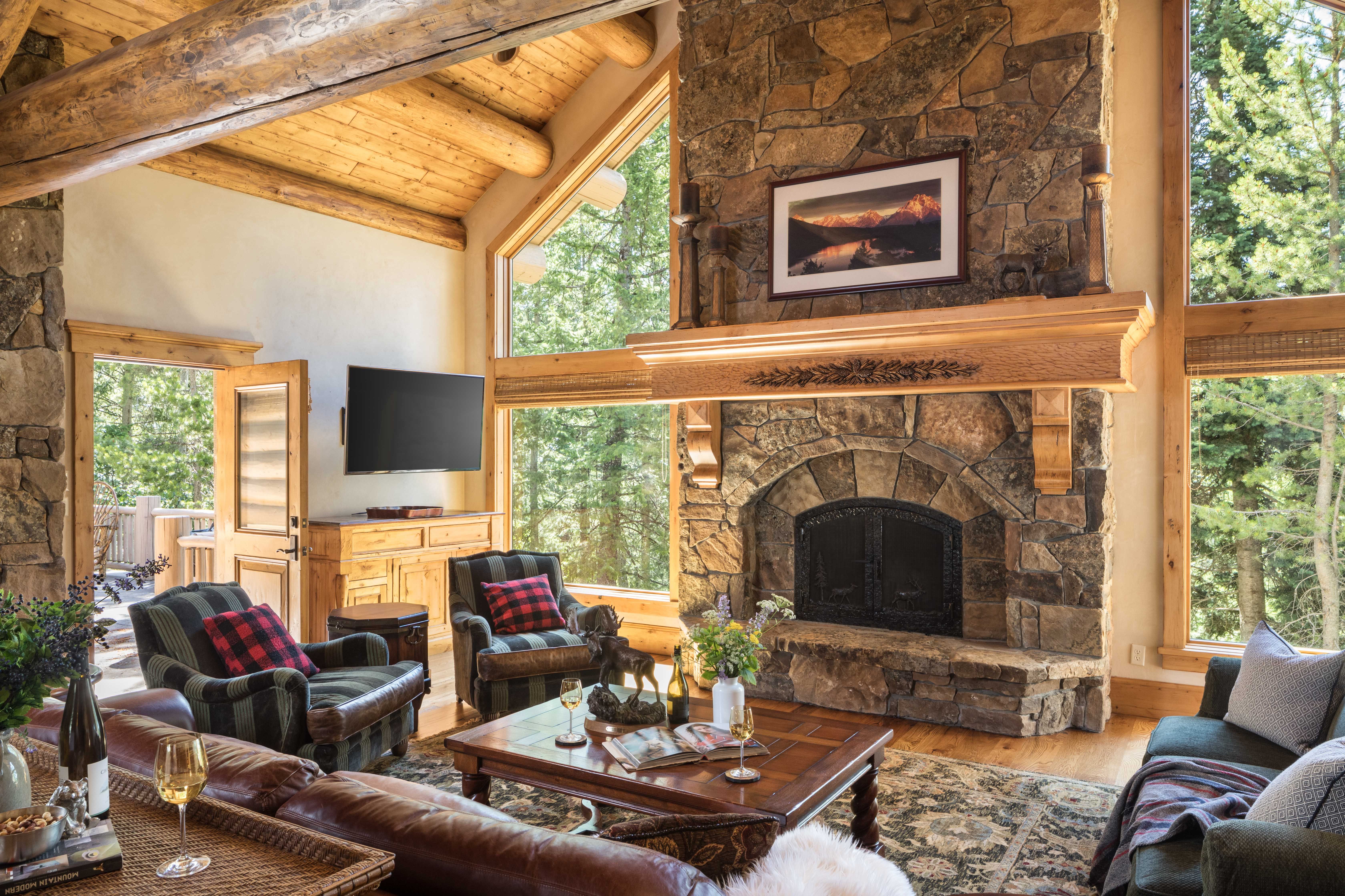 Rustic mountain lodge living room with vaulted wood ceilings, a large stone fireplace, cozy seating with plaid accents, and tall windows framing a forested view.