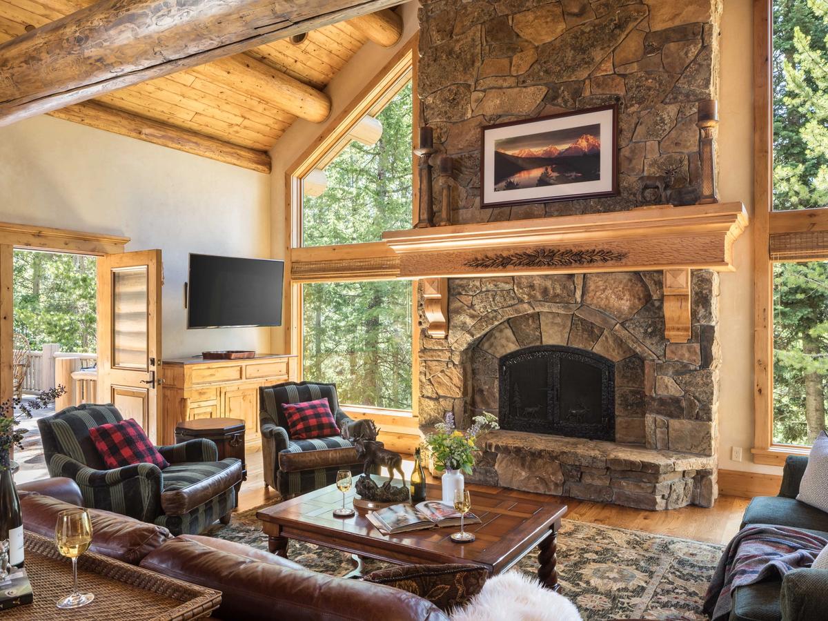 Rustic mountain lodge living room with vaulted wood ceilings, a large stone fireplace, cozy seating with plaid accents, and tall windows framing a forested view.