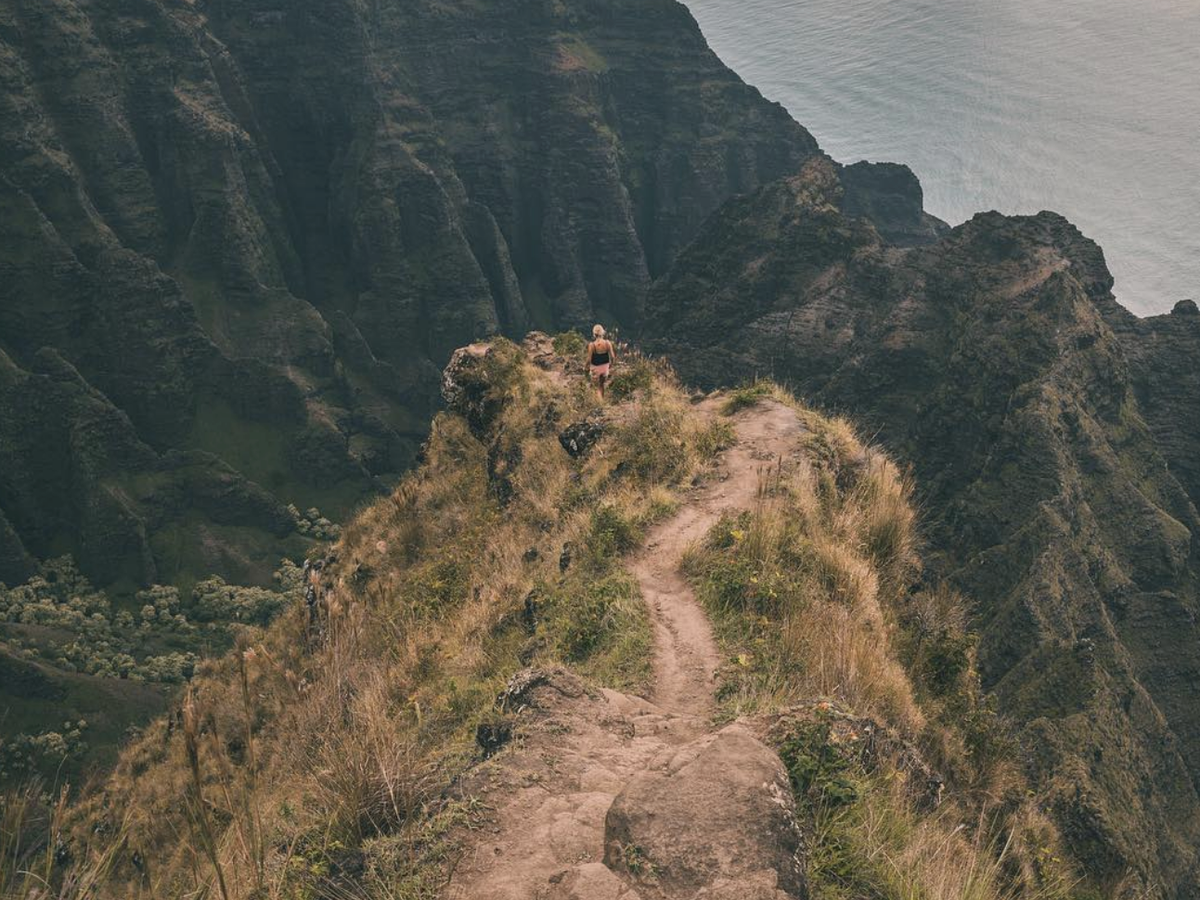 A lone hiker walks along a narrow cliffside trail overlooking the dramatic ridges and valleys of Kauai’s Nā Pali Coast.
