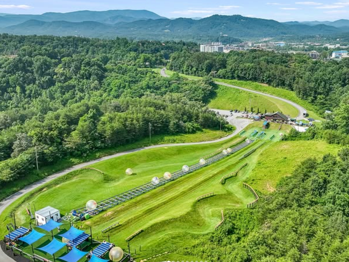 An aerial view shows rolling green hills with zorb balls on winding tracks, set against a backdrop of forest and mountains.