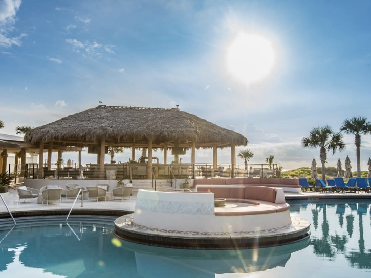 A luxurious resort pool area with a thatched-roof tiki bar, sun loungers, and palm trees under a bright blue sky. The pool features a circular built-in seating area with a fire pit, creating a relaxing tropical atmosphere