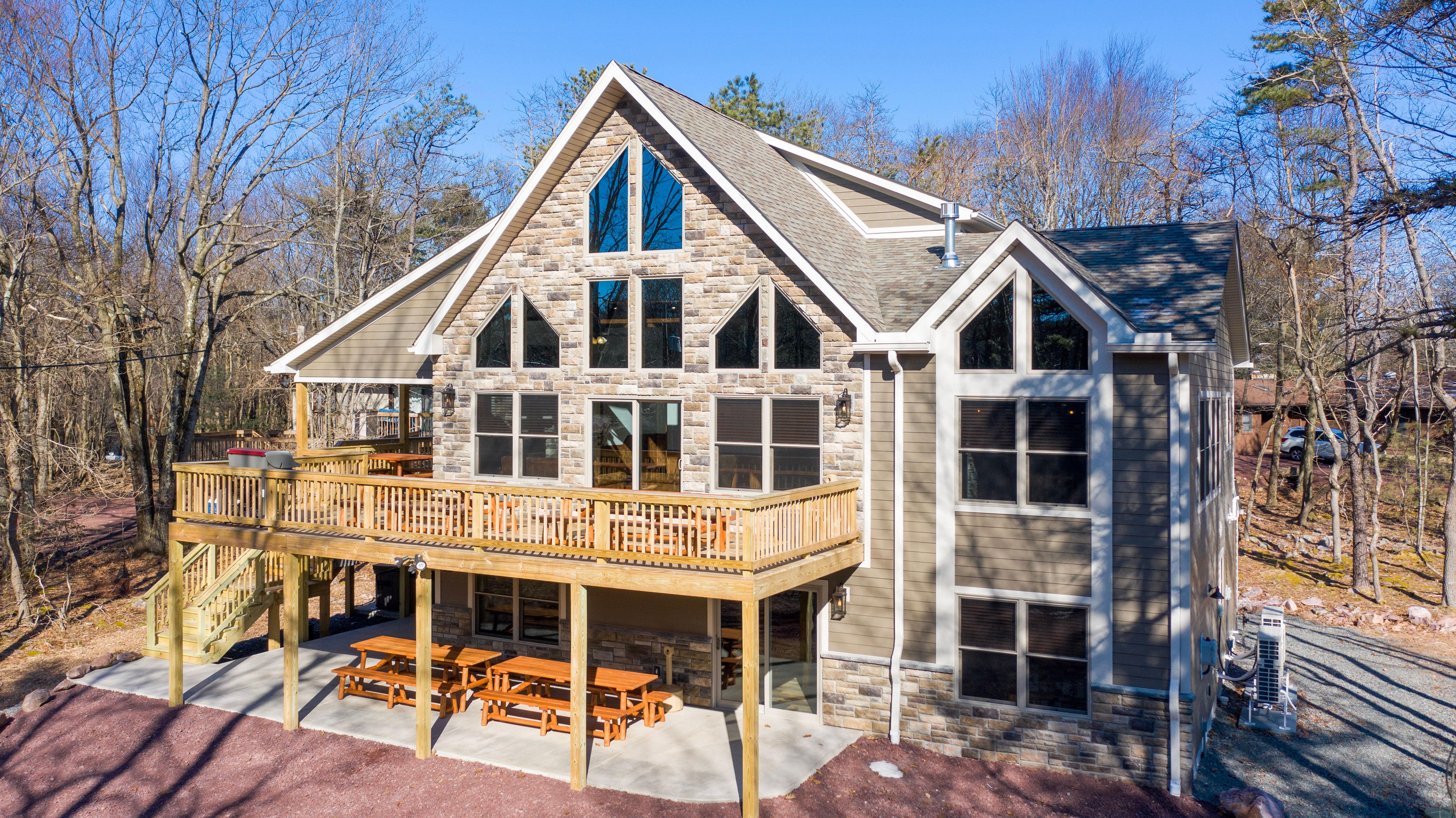 Charming two-story cabin with a stone and wood exterior, large triangular windows, an upper-level wooden deck, and picnic tables on a patio beneath, surrounded by trees.