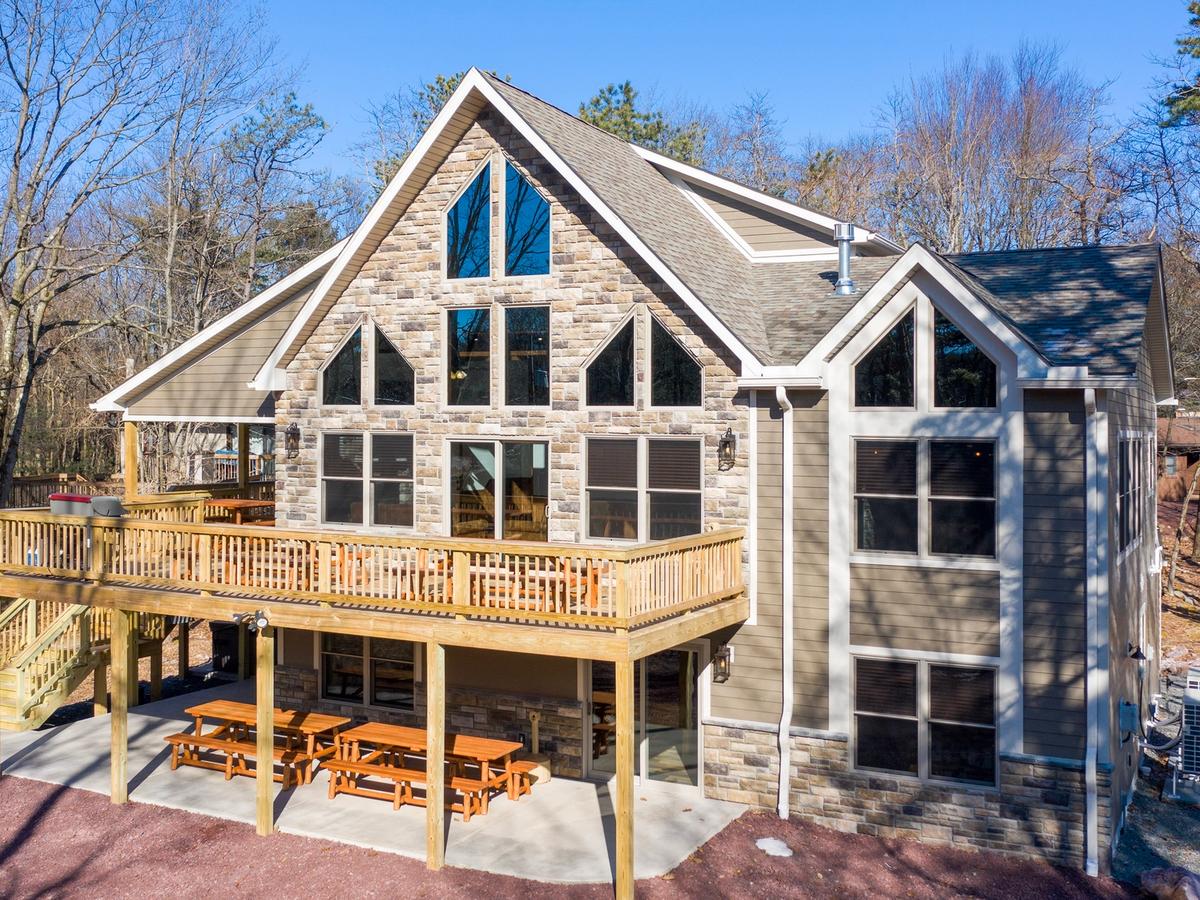 Charming two-story cabin with a stone and wood exterior, large triangular windows, an upper-level wooden deck, and picnic tables on a patio beneath, surrounded by trees.