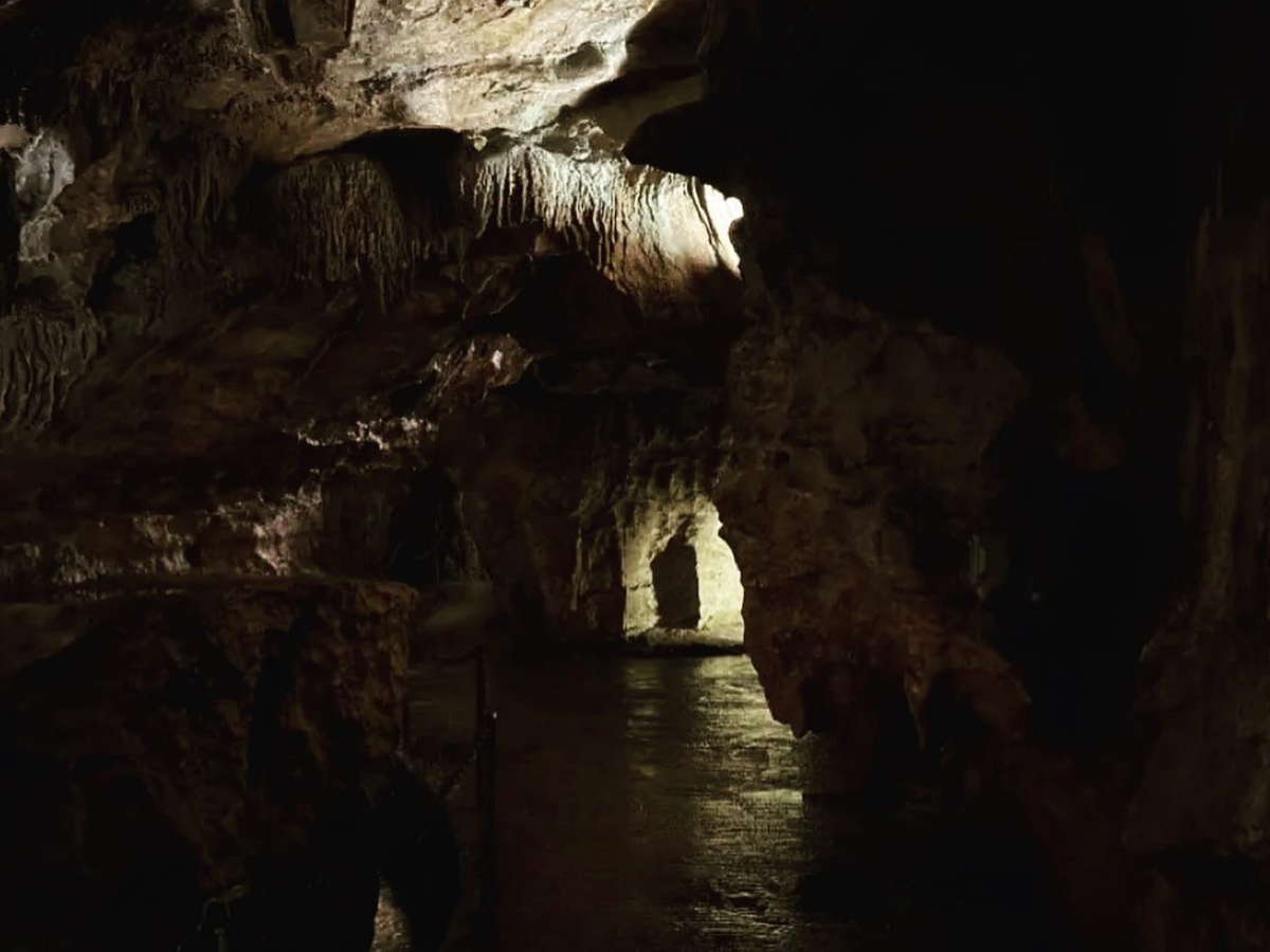 Dimly lit cave interior with rough rock walls, hanging formations, and a glowing passageway illuminated by soft artificial light.