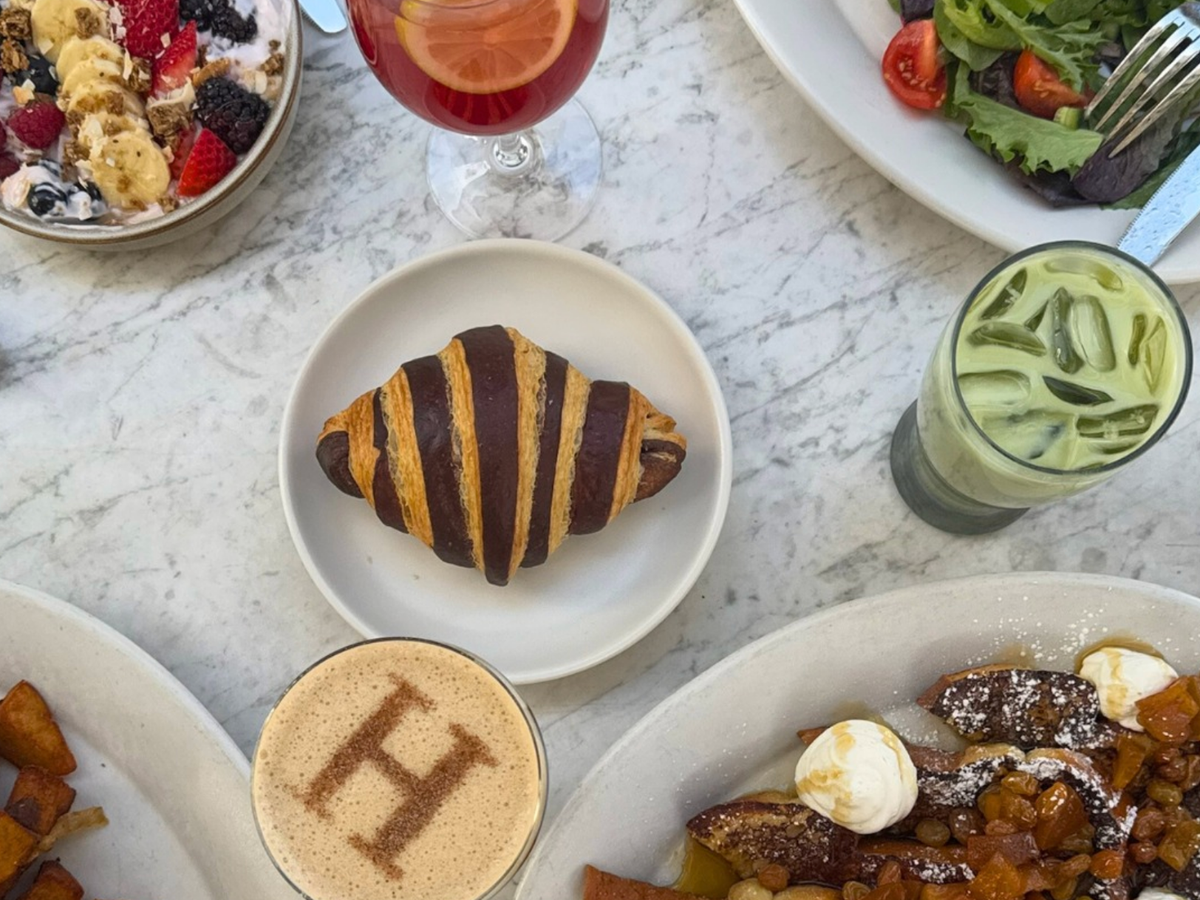 A marble table set with a chocolate-striped croissant, French toast topped with fruit and whipped cream, a yogurt bowl with bananas and berries, a salad plate, and drinks including iced matcha, sangria, and a latte with a cinnamon "H" design.