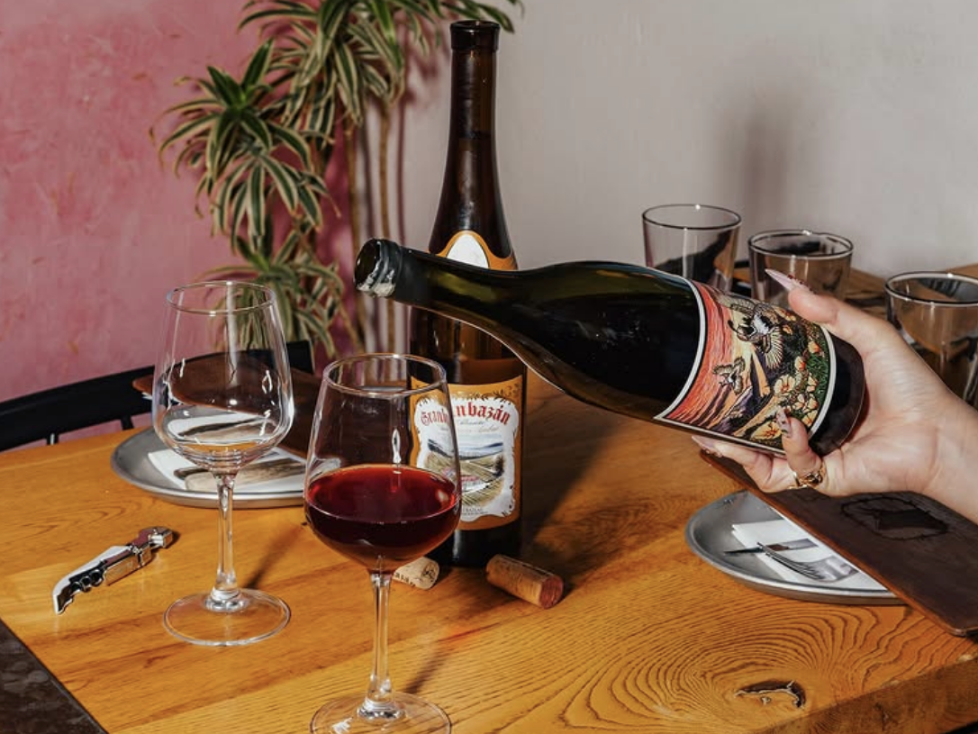 Hand pouring red wine into a glass at a wooden table set with wine glasses, bottles, corks, and casual place settings, with a pink accent wall and potted plant in the background.