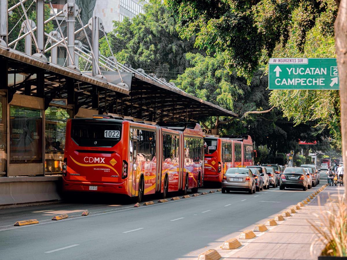 Bright red CDMX public buses lined up at a Mexico City bus station with cars and trees nearby. A common way travelers connect before flying to Huatulco.