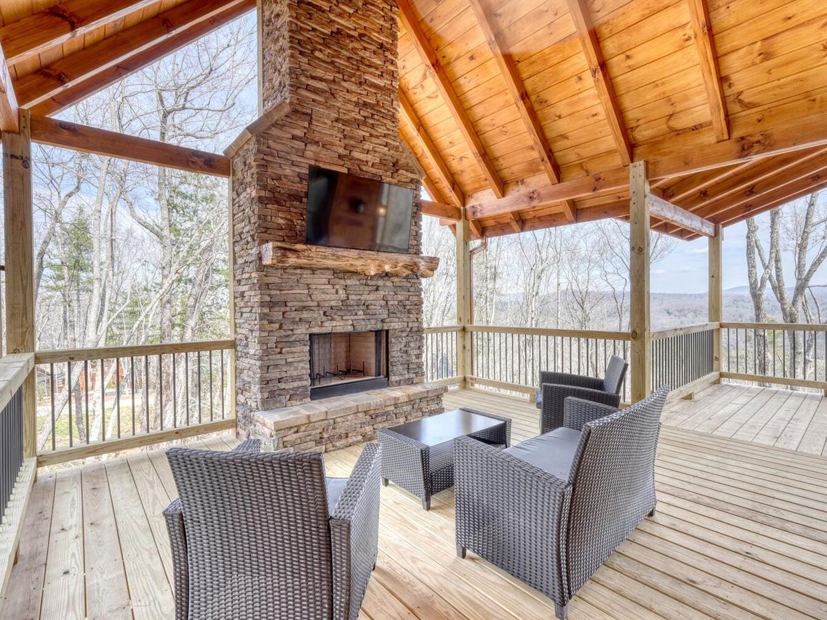 Covered deck with a big stone fireplace, wicker seating, and sweeping views of the woods and sky. This cabin is built for large groups who want comfort, space, and a hot tub close by.