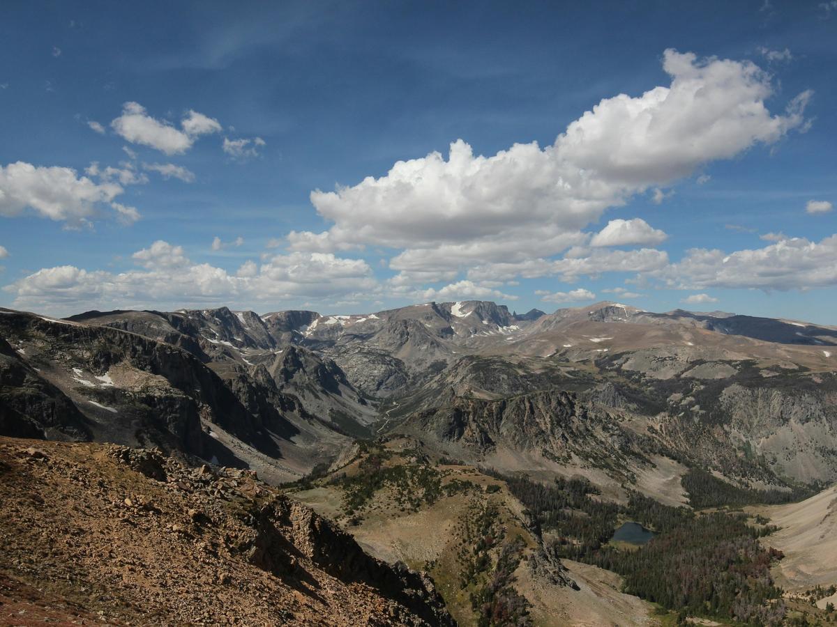 Expansive mountain landscape along the Beartooth Highway, featuring rugged peaks, deep valleys, and alpine lakes under a bright blue sky with scattered clouds