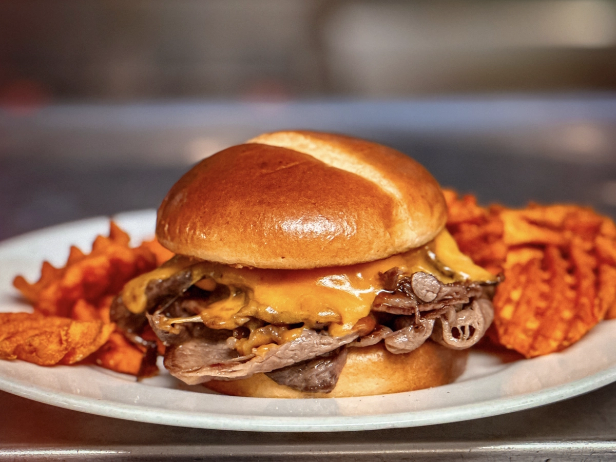 Close-up of a beef and cheddar sandwich on a toasted bun, served with crispy waffle fries on a white plate.
