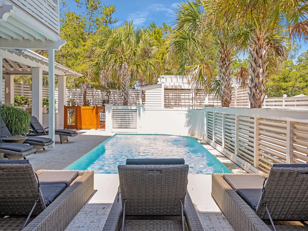 A private, sun-drenched backyard pool area lined with wicker loungers, tropical palm trees, and modern white fencing, perfect for relaxing in a serene coastal setting.