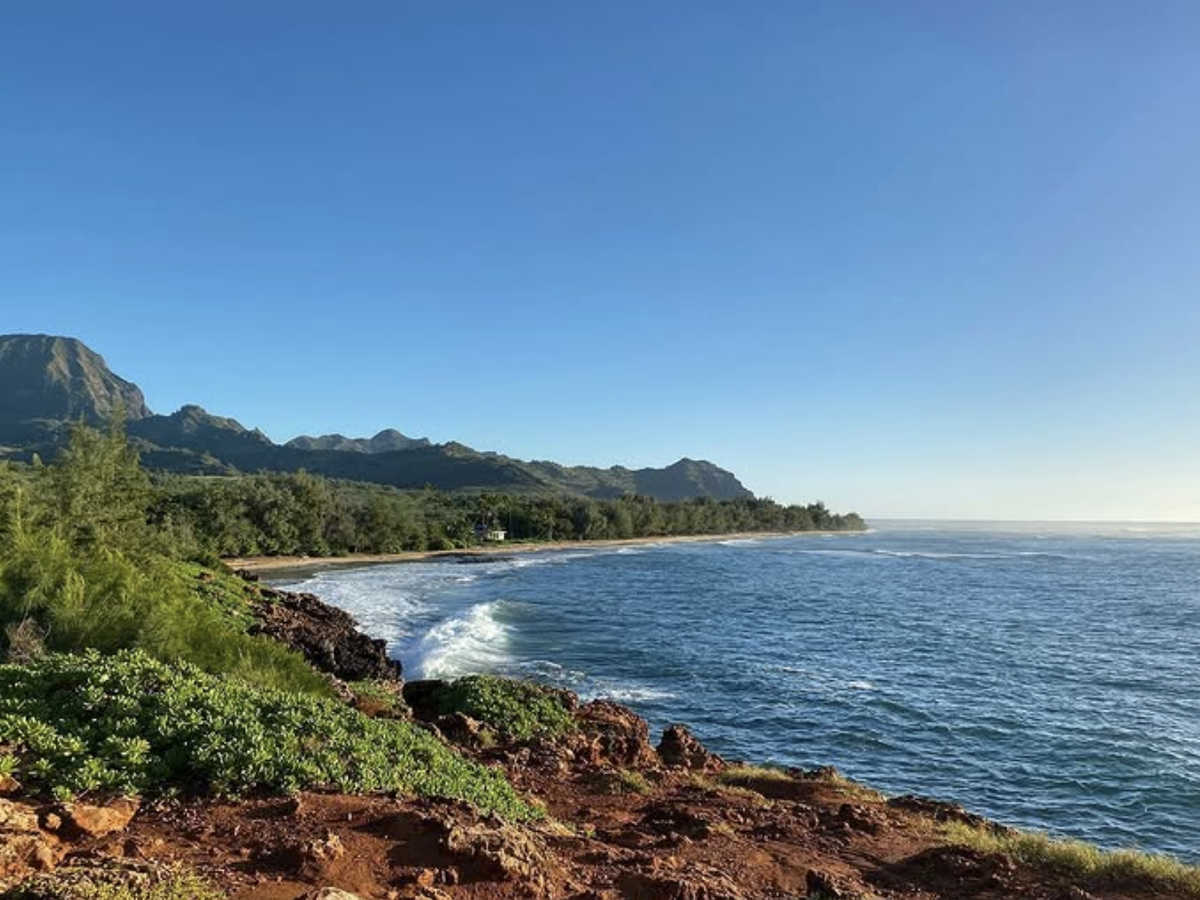 A clear morning view of Kauai’s rugged red cliffs and lush greenery overlooking a calm blue ocean with mountains in the distance.