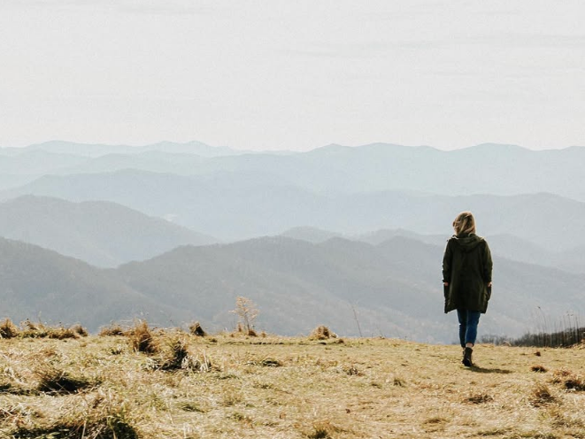 A solitary hiker in a hooded jacket walks along a grassy ridge, overlooking rolling mountain peaks that fade into the misty horizon. The scene exudes tranquility and adventure