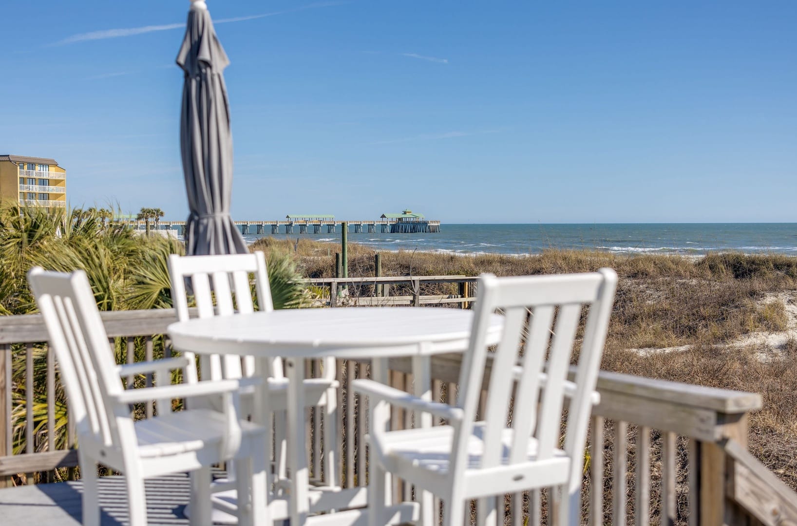 A white outdoor table and chairs sit on a beachfront deck with clear views of the ocean and a nearby pier. This quiet seating area is perfect for morning coffee or sunset drinks by the shore. It captures the relaxed coastal feel of staying right on the beach.