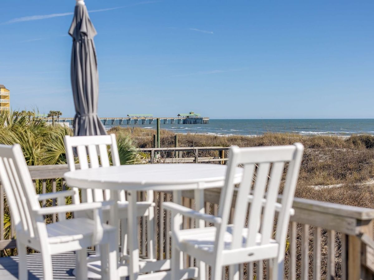 A white outdoor table and chairs sit on a beachfront deck with clear views of the ocean and a nearby pier. This quiet seating area is perfect for morning coffee or sunset drinks by the shore. It captures the relaxed coastal feel of staying right on the beach.