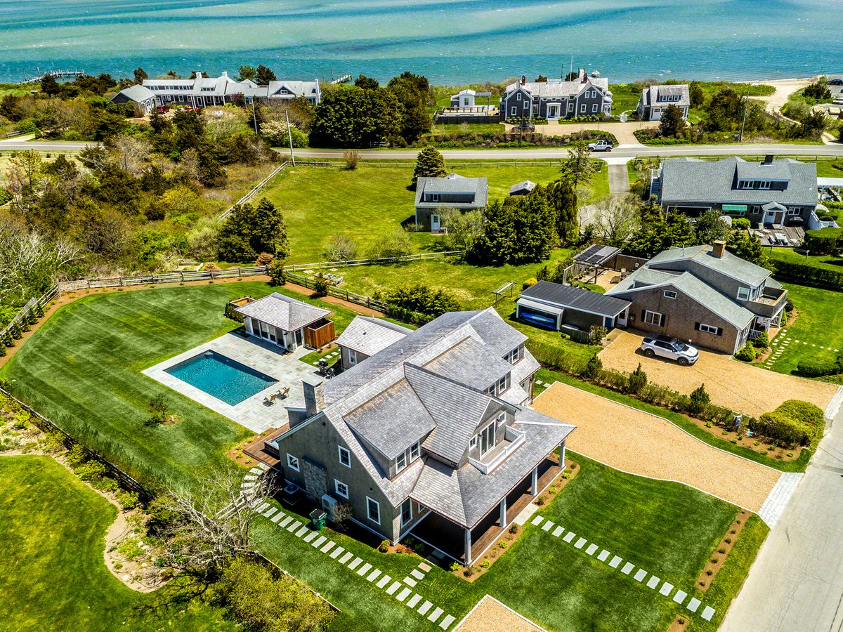An aerial view of a luxury coastal property with a pool, guesthouse, manicured lawn, and ocean views, located in an upscale seaside neighborhood.