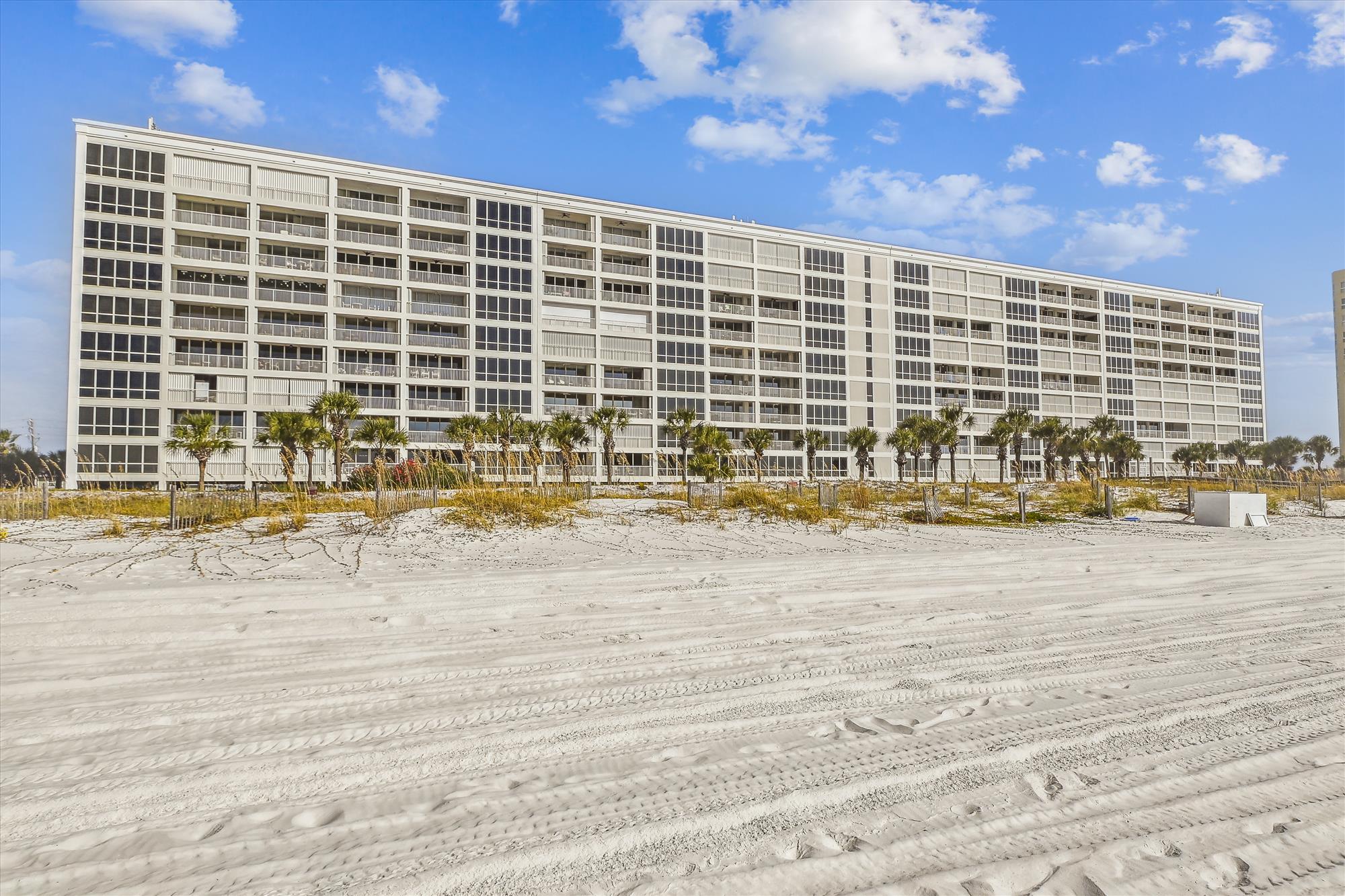 A wide beachfront condominium complex with large glass-enclosed balconies, framed by palm trees and white sand under a bright blue sky.