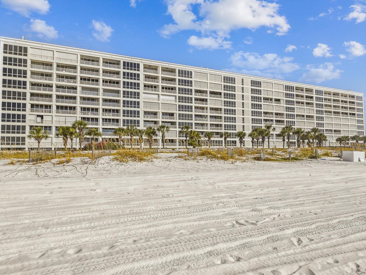 A wide beachfront condominium complex with large glass-enclosed balconies, framed by palm trees and white sand under a bright blue sky.