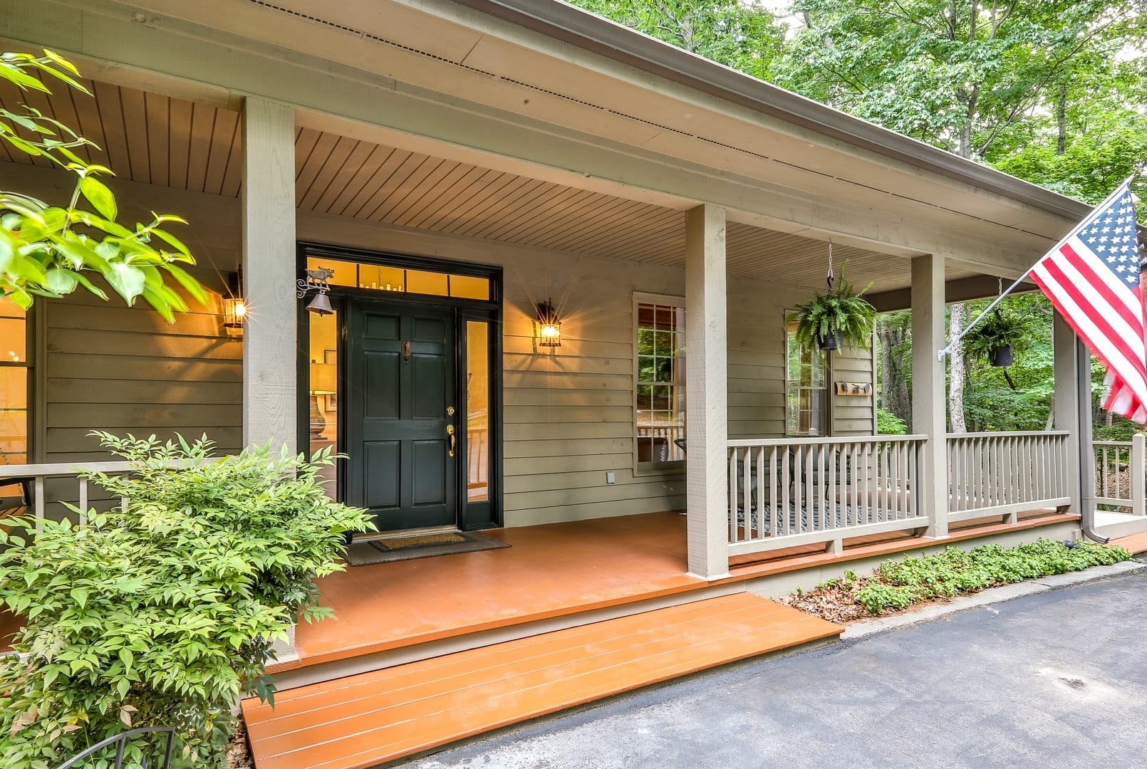 Inviting front porch with a dark green door, hanging ferns, warm lighting, and an American flag, surrounded by trees and greenery.