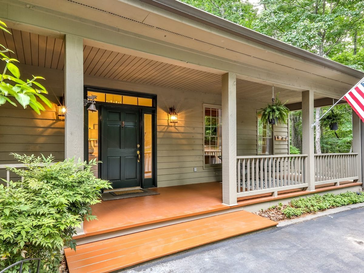 Inviting front porch with a dark green door, hanging ferns, warm lighting, and an American flag, surrounded by trees and greenery.