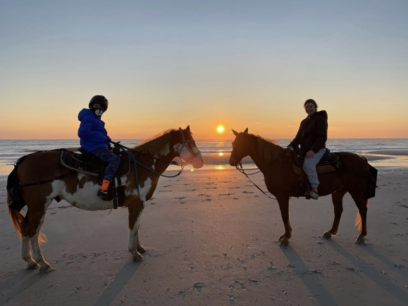 Two horseback riders, a child in a blue jacket and helmet and an adult in a dark coat, enjoy a peaceful sunset ride along a sandy beach. The sun sets over the ocean, casting a golden glow on the water and creating long shadows on the shore