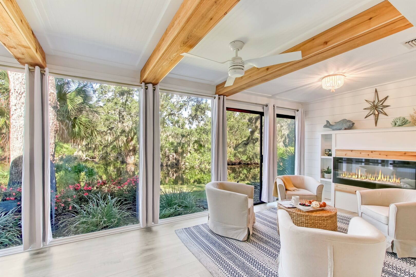 A serene sunroom with floor-to-ceiling windows offering a lush lagoon view. The space features cozy white armchairs, a woven coffee table, and a modern linear fireplace set against shiplap walls. Exposed wooden beams and soft natural light enhance the tranquil, coastal ambiance