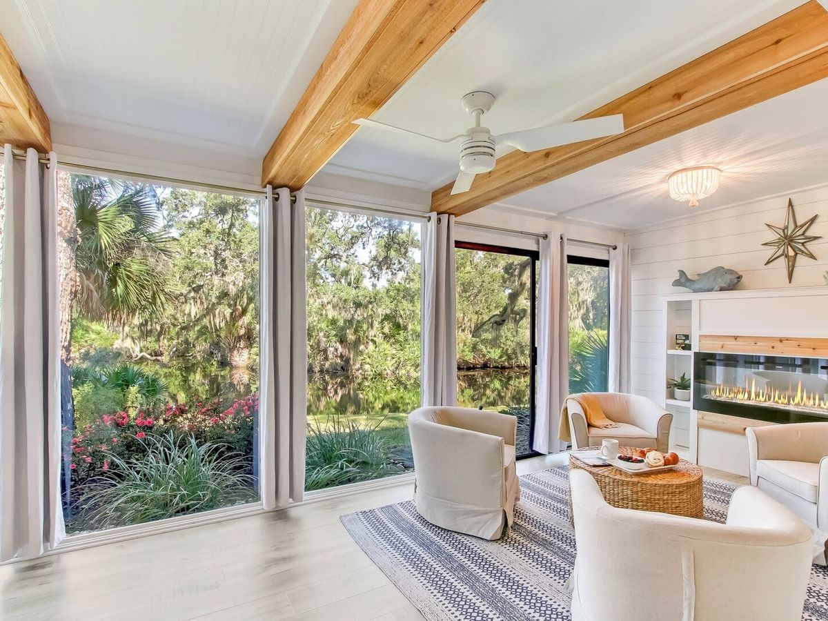 A serene sunroom with floor-to-ceiling windows offering a lush lagoon view. The space features cozy white armchairs, a woven coffee table, and a modern linear fireplace set against shiplap walls. Exposed wooden beams and soft natural light enhance the tranquil, coastal ambiance
