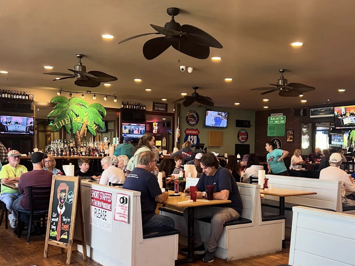 A lively, casual restaurant interior with white booths, ceiling fans, and tropical-themed bar décor featuring a large "The Peach" sign; guests are dining and watching sports on multiple TVs.