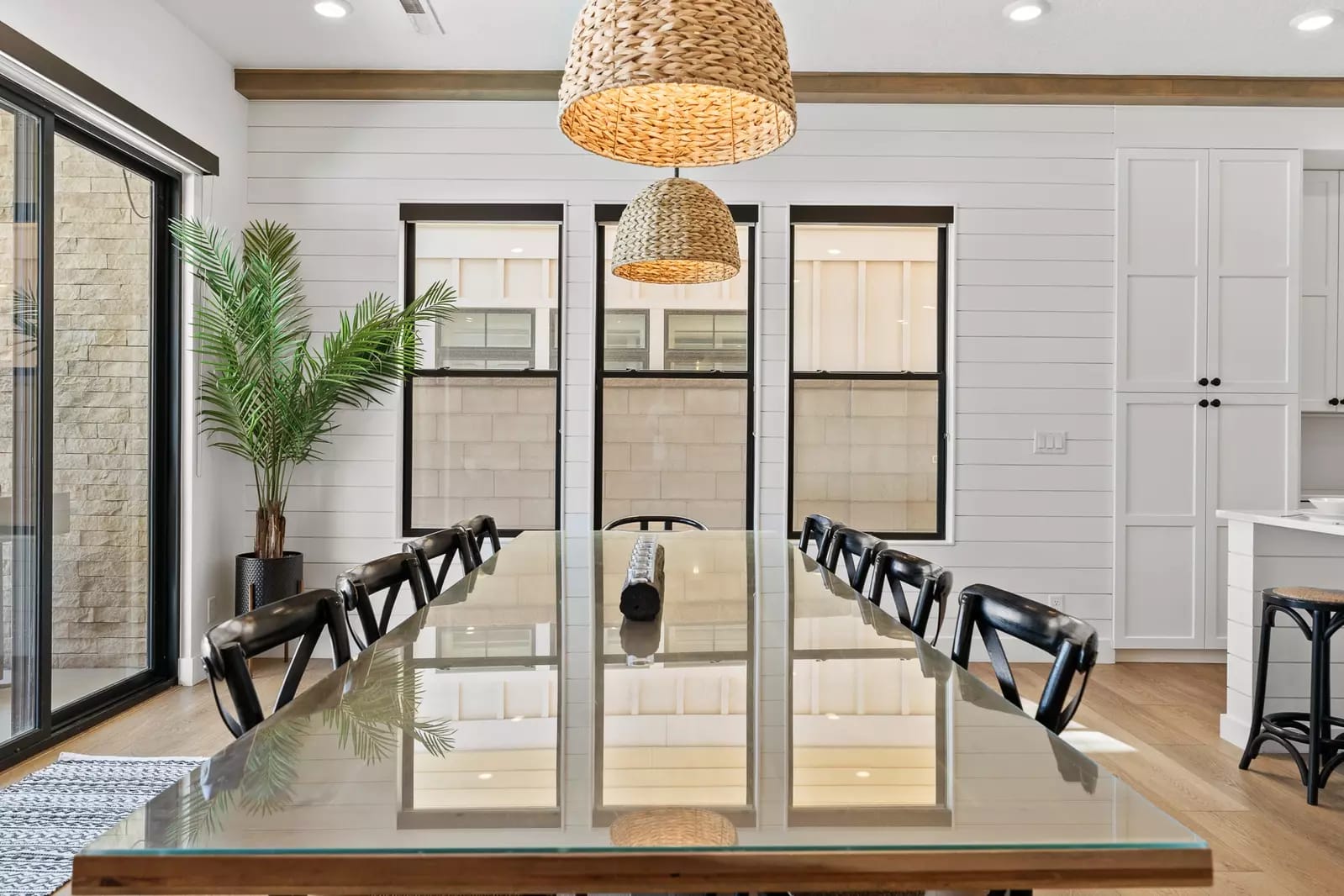Bright coastal dining room with a long glass table, black chairs, woven pendant lights, white shiplap walls, and large windows letting in natural light.