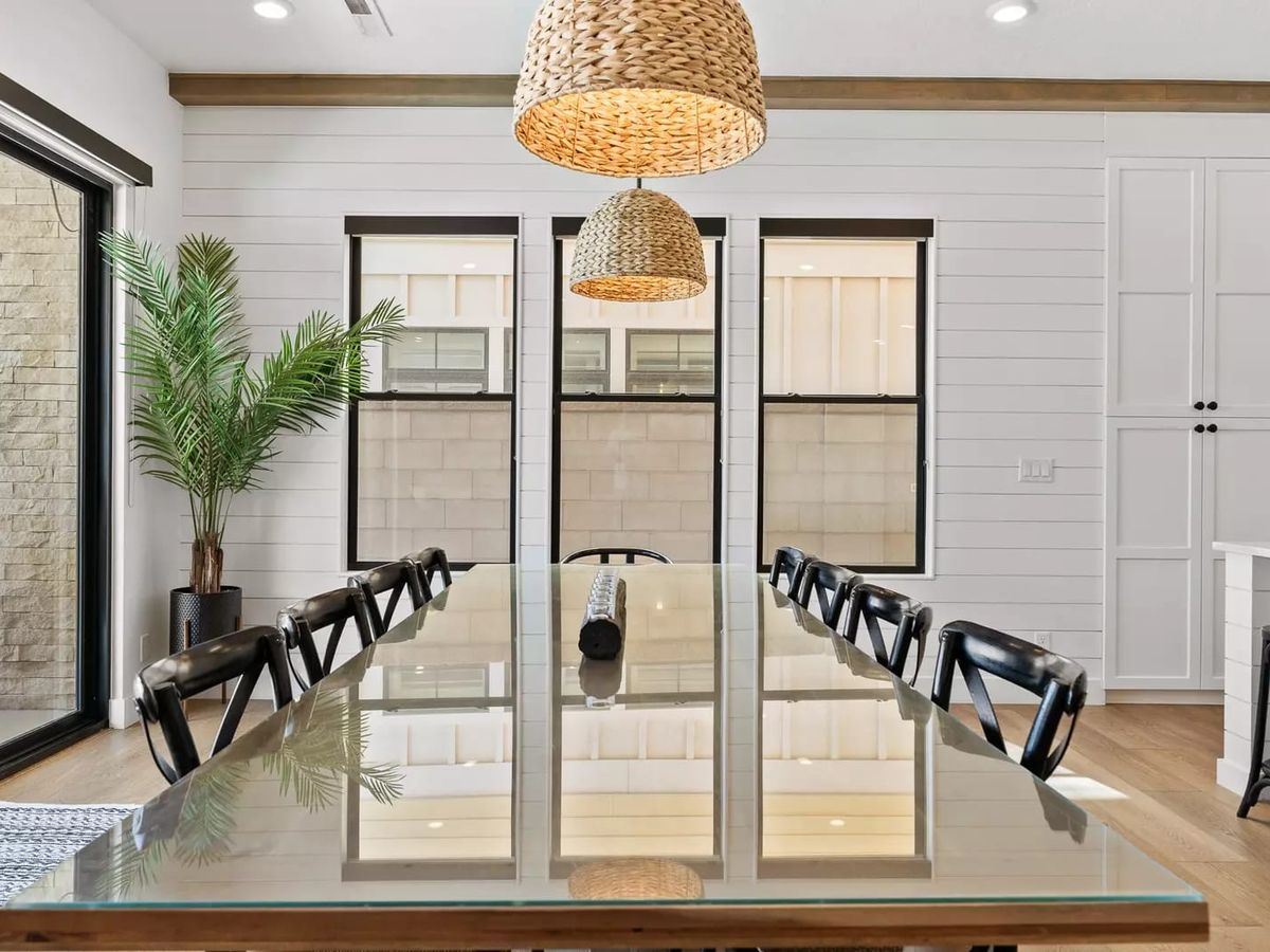 Bright coastal dining room with a long glass table, black chairs, woven pendant lights, white shiplap walls, and large windows letting in natural light.