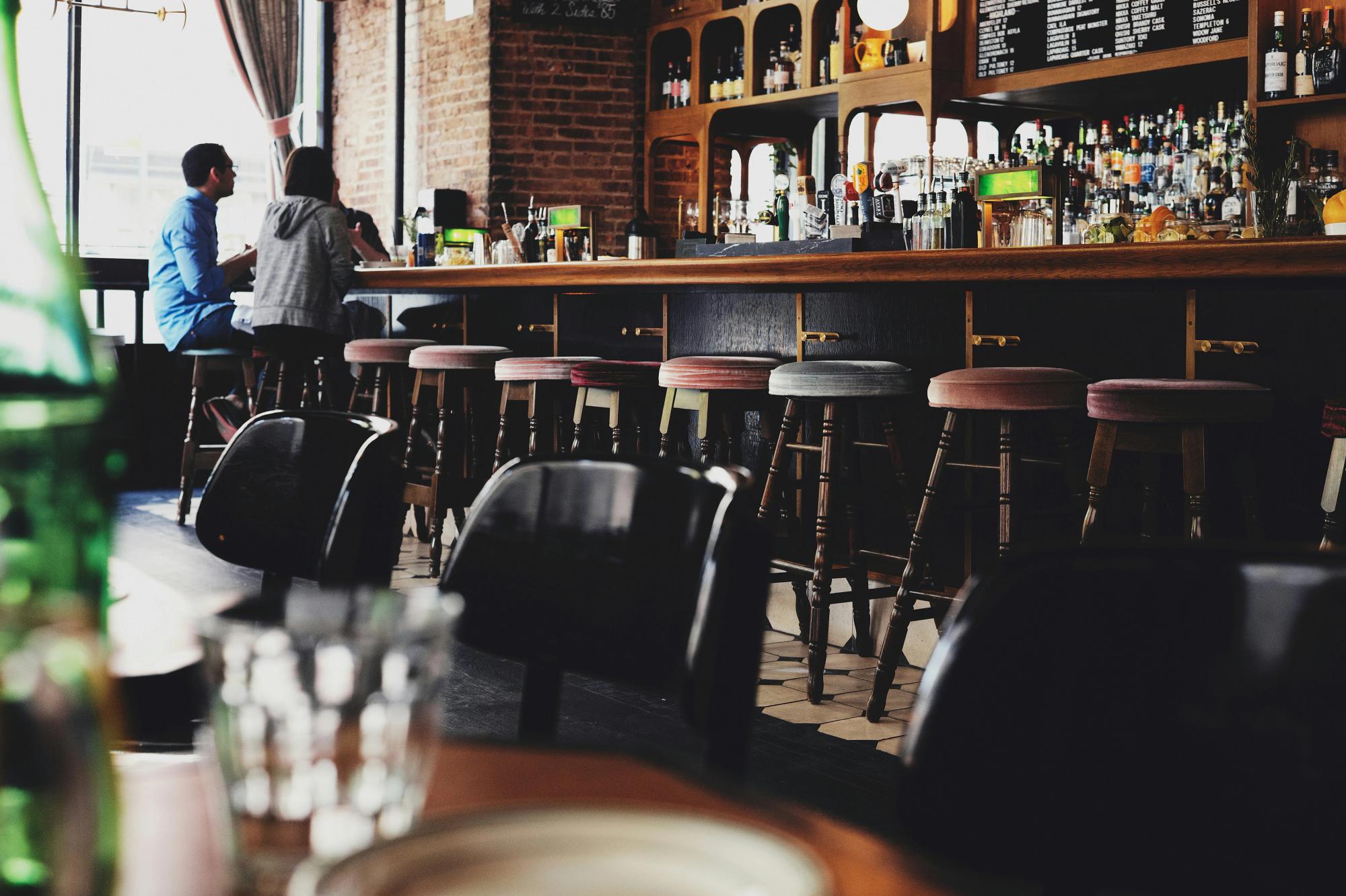 A cozy, stylish bar with wooden stools, a well-stocked back bar, and a couple seated at the counter in conversation.