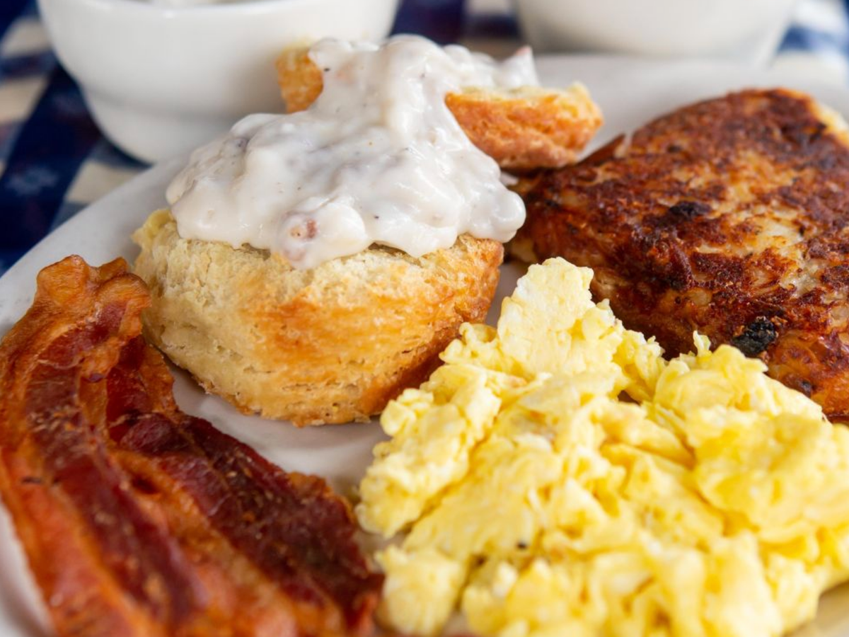 A classic Southern breakfast plate with crispy bacon, scrambled eggs, a biscuit topped with sausage gravy, and golden hash browns.