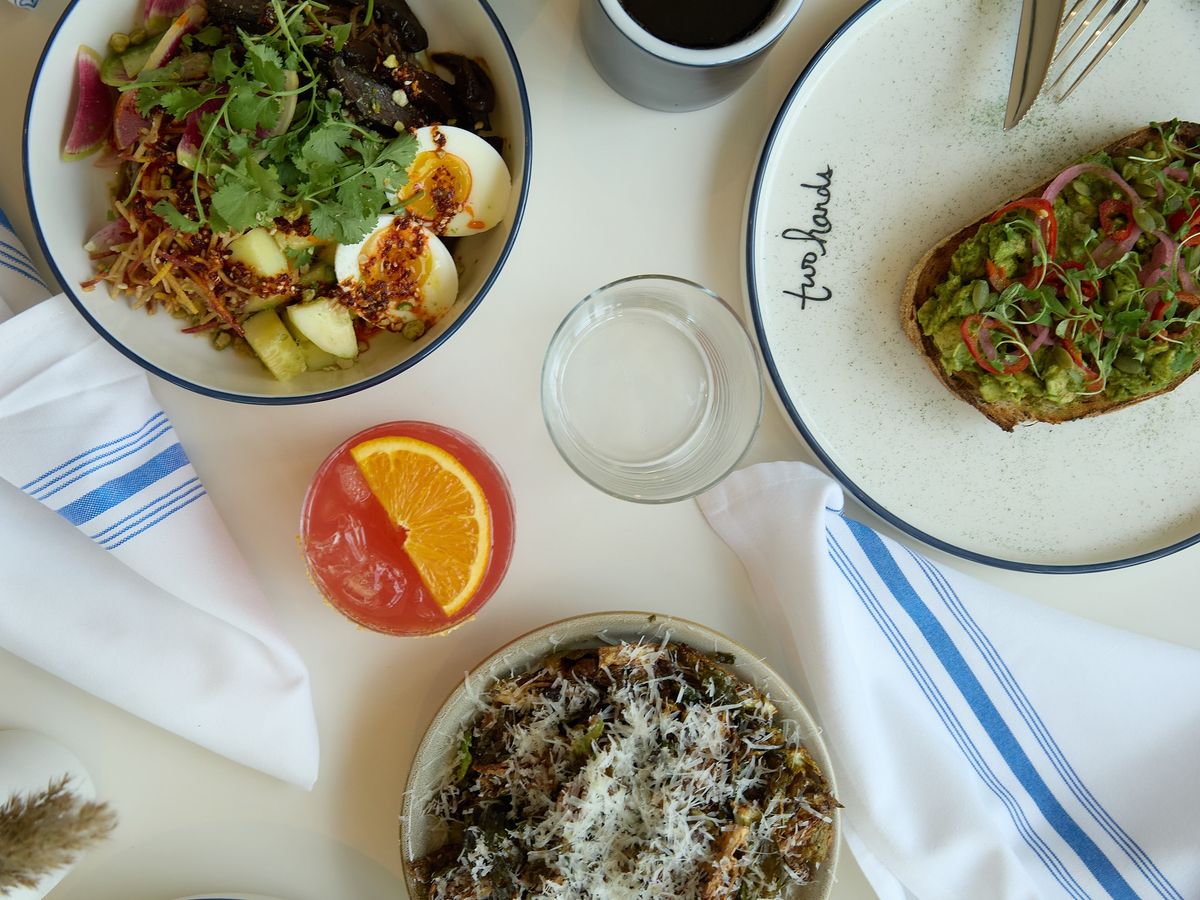 A fresh and vibrant café spread featuring avocado toast, colorful salads, and refreshing drinks. The clean table setup with striped napkins and natural light captures the relaxed energy of a modern brunch spo