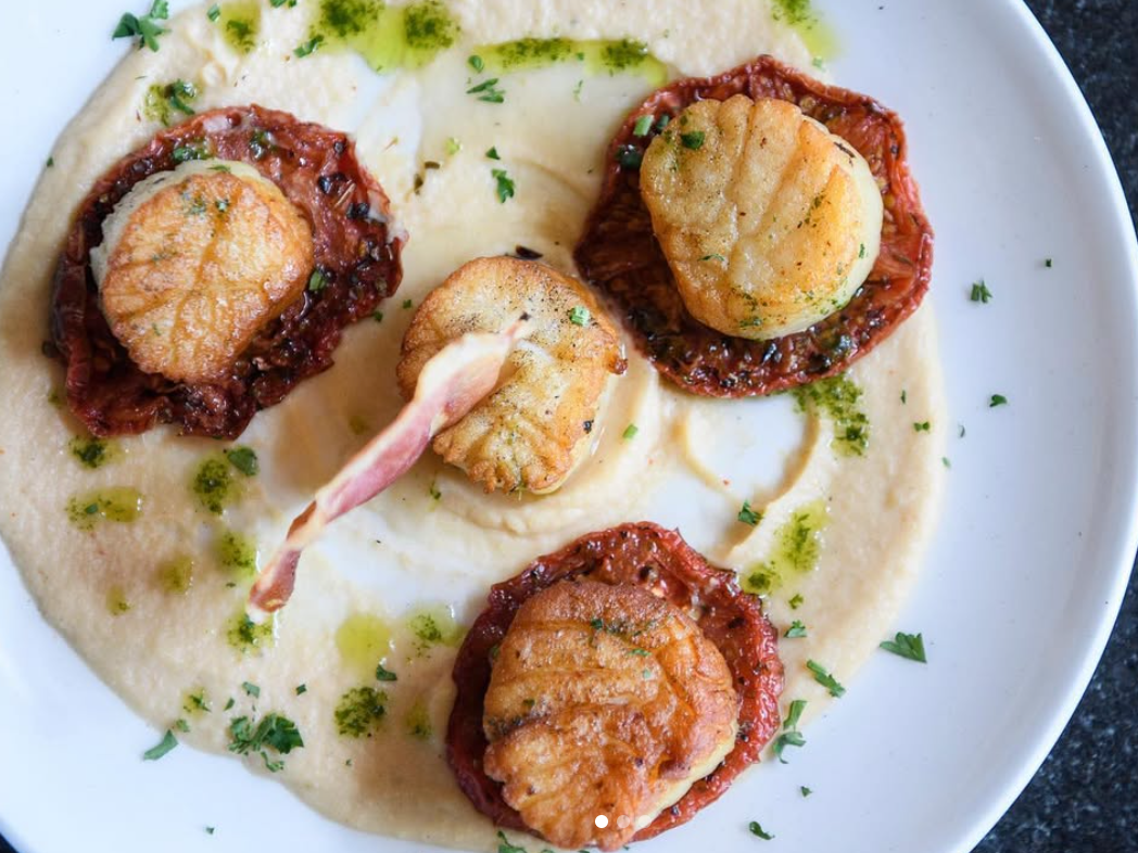 Four perfectly seared scallops sit atop roasted tomatoes on a creamy purée, garnished with herbs and a drizzle of green sauce