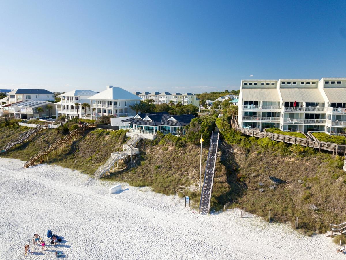 Aerial view of beachfront condos and houses with staircases leading down a sandy dune to a bright white beach, where a few people are sitting near the shoreline under a clear blue sky.